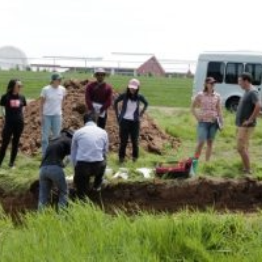 Field trip photo of students and instructor standing in front of a soil pit. In the background are farm buildings and the bus.