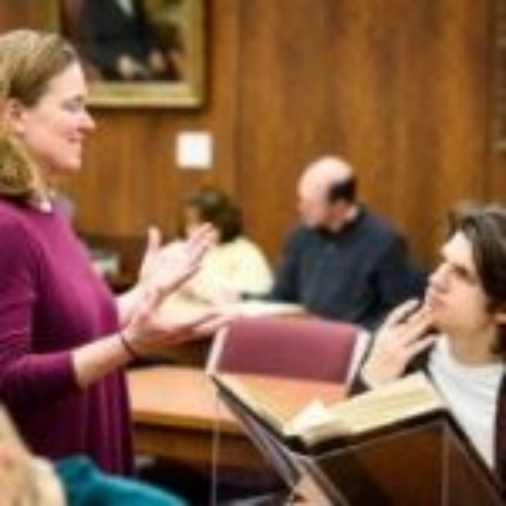 Adult female standing in front of a student holding an open book as they discuss a topic. In background are other individuals seated at tables. 
