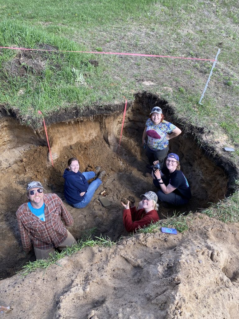 Students standing and sitting in a soil pit.